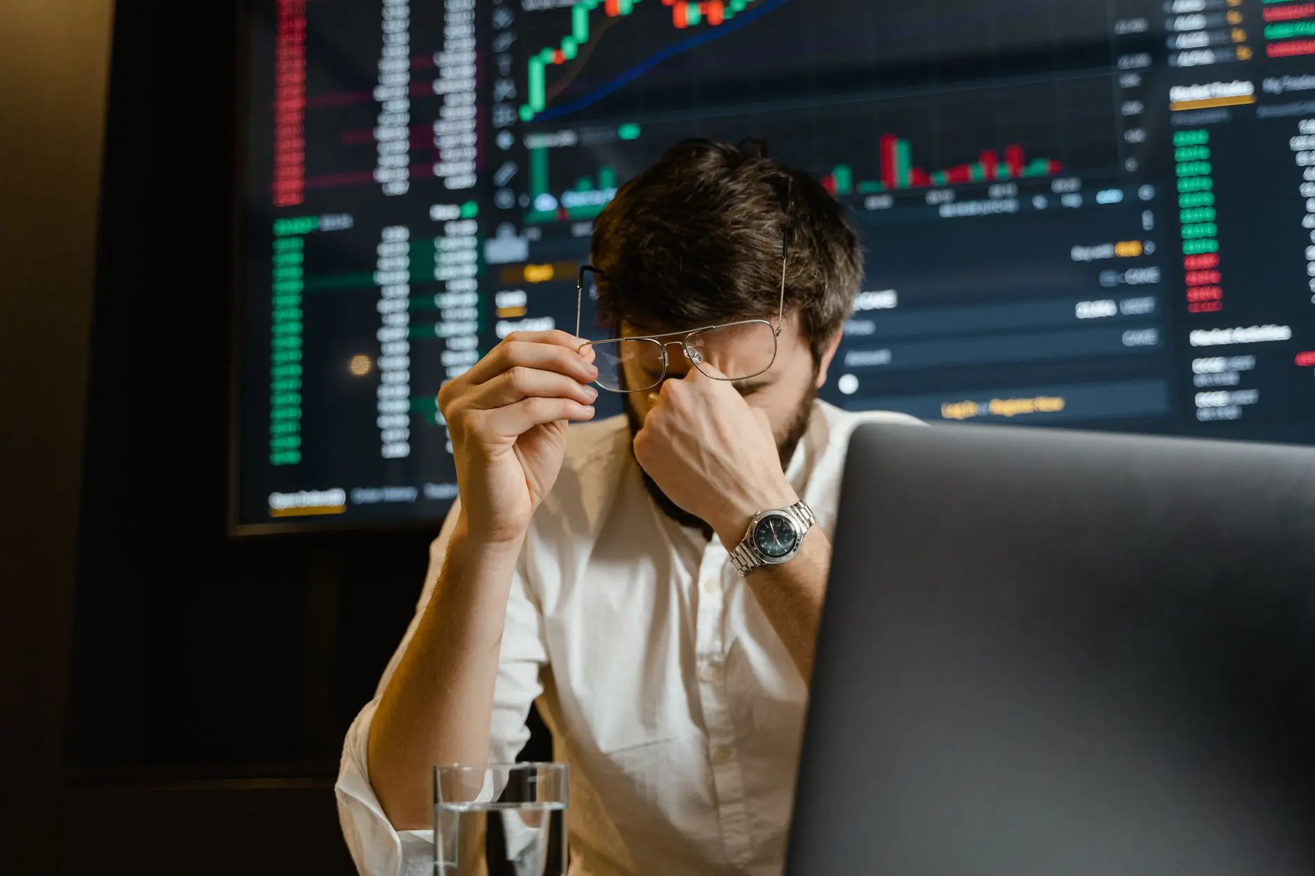 A stressed trader holding their glasses, looking at stock market data on a screen.