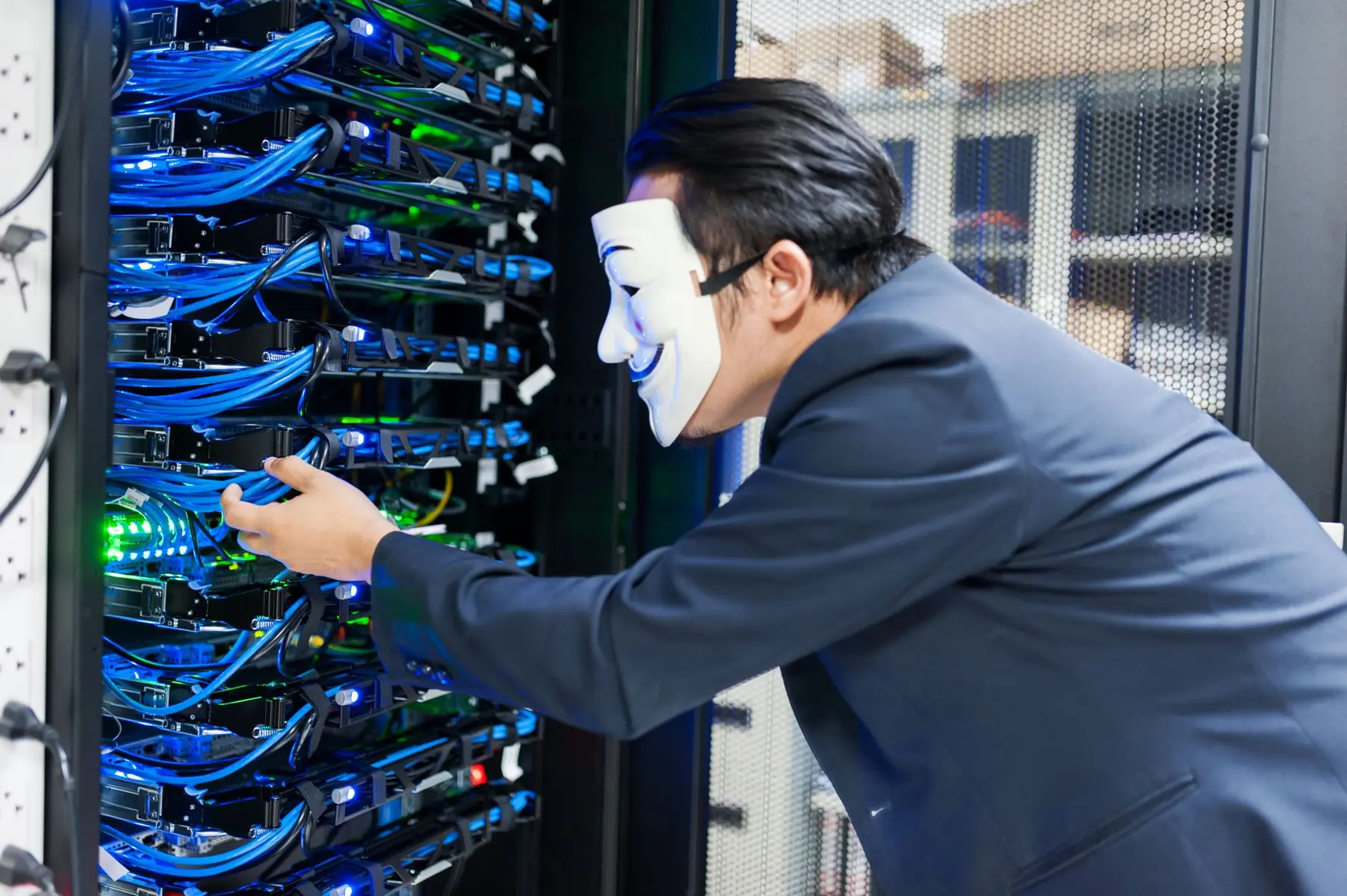 A person in a business suit wearing a white Guy Fawkes mask interacting with server racks in a data center
