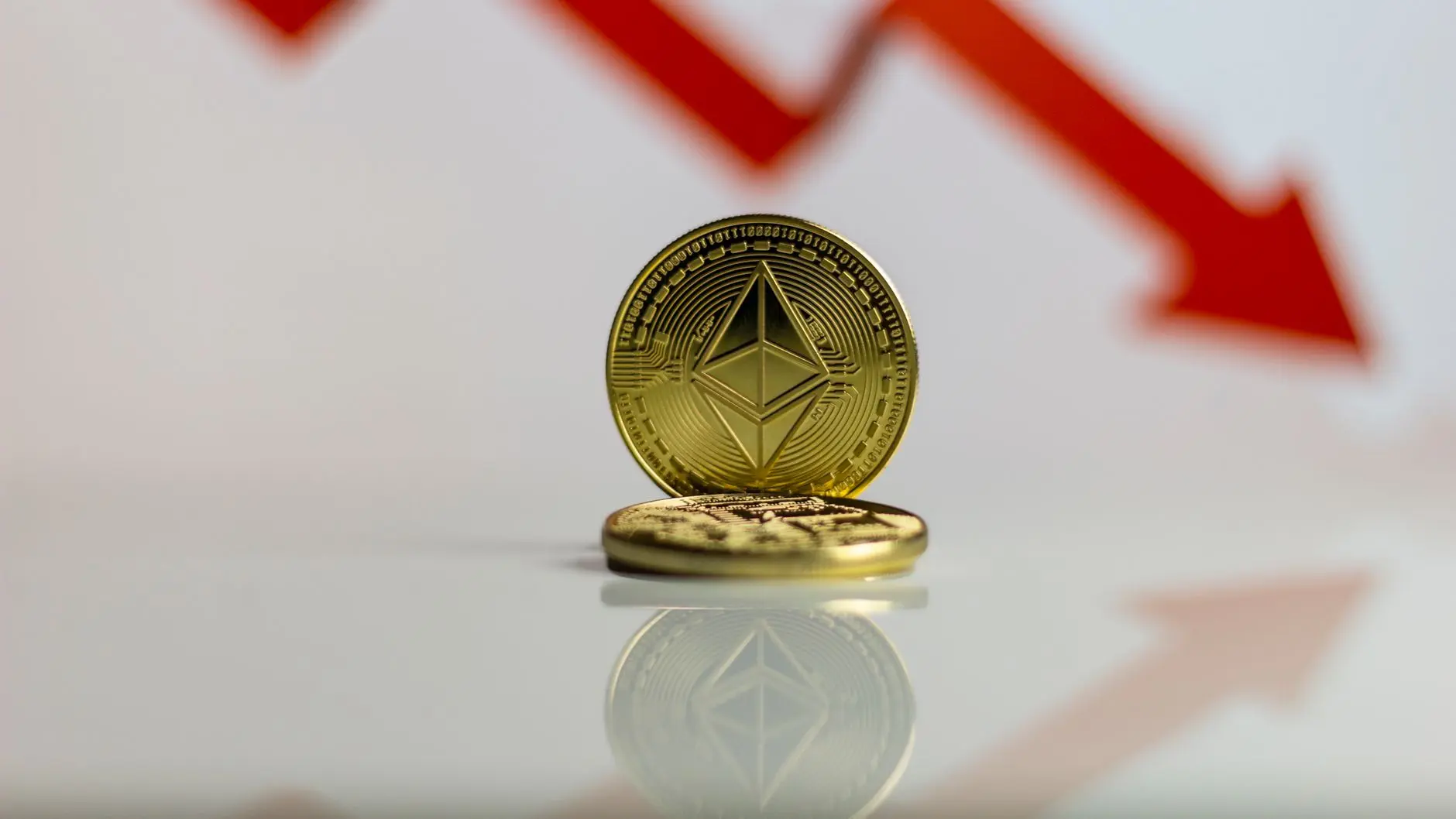 Physical gold Ethereum coins stacked in front of a blurred red downward trending arrow graph