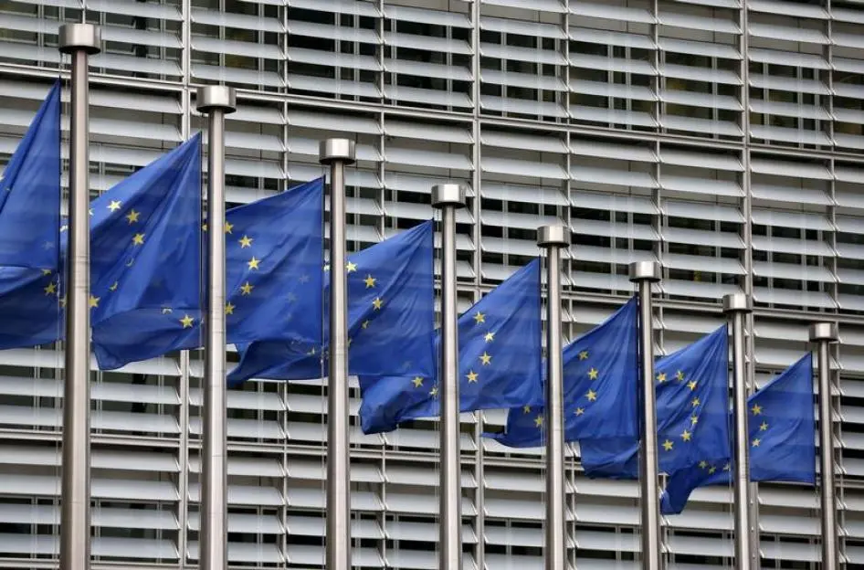 A row of European Union flags flying in front of a modern glass building with horizontal louvers