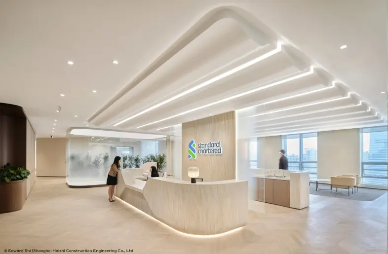 Modern interior of a Standard Chartered bank office with a light wood reception desk and glowing ceiling lights.