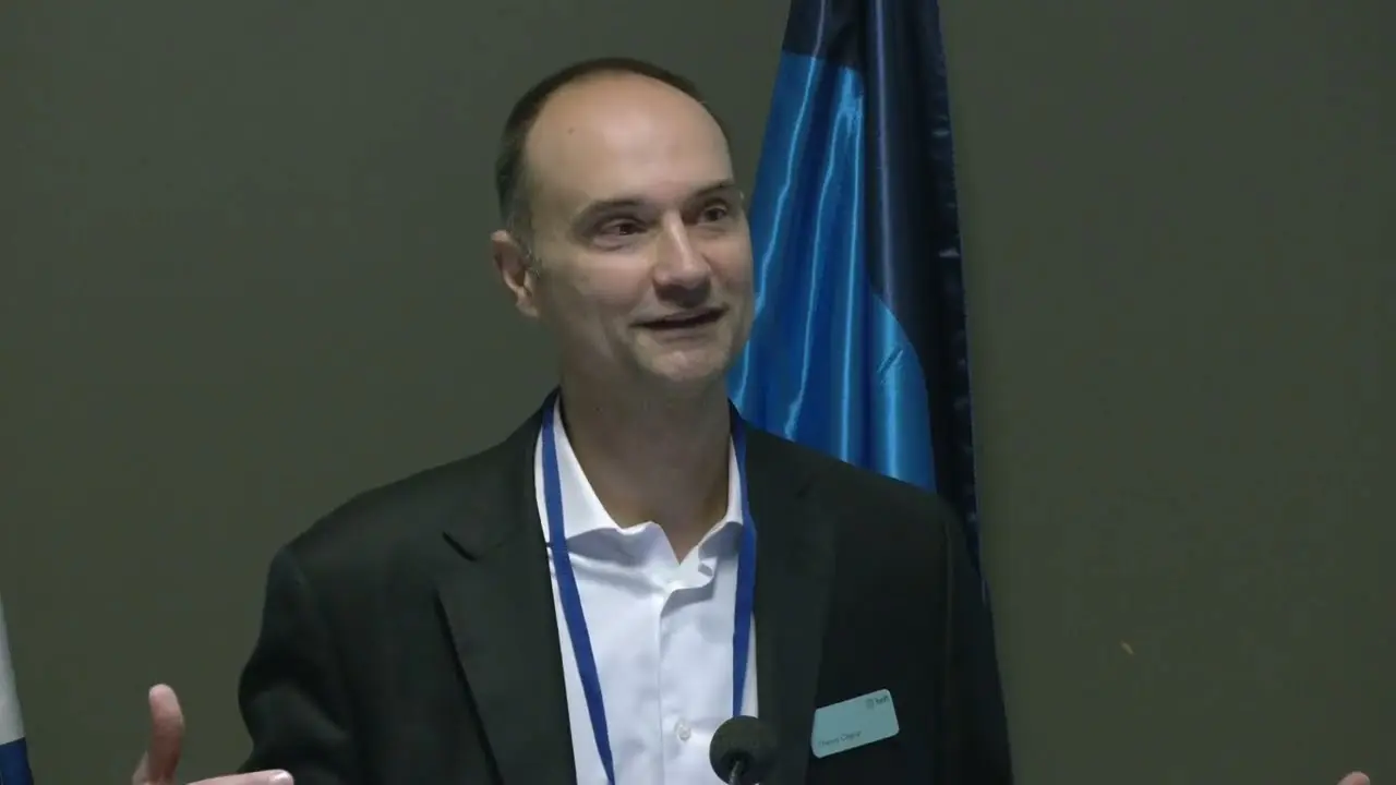 Thierry Chilosi speaking at an event with flags in the background.