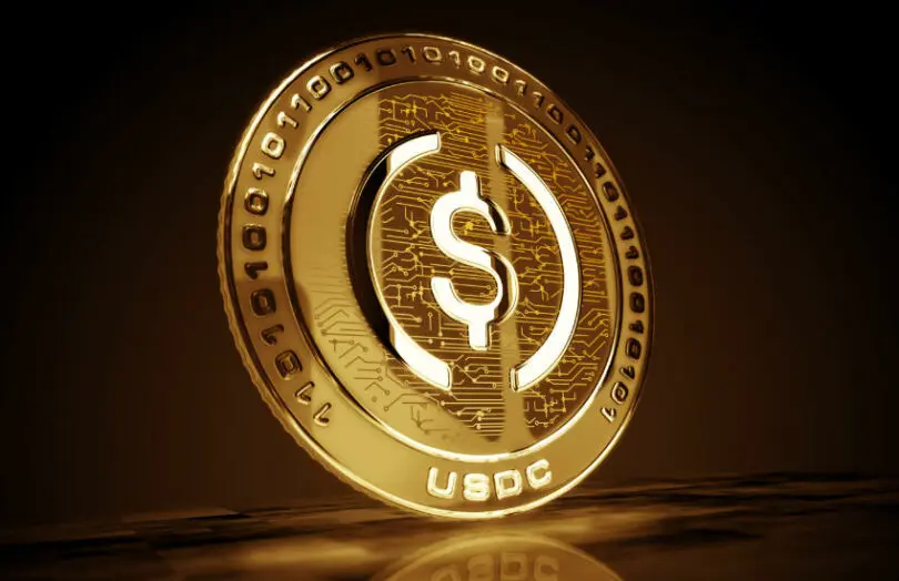 A gold 3D digital coin featuring a dollar sign and the text USDC on a dark reflective surface.