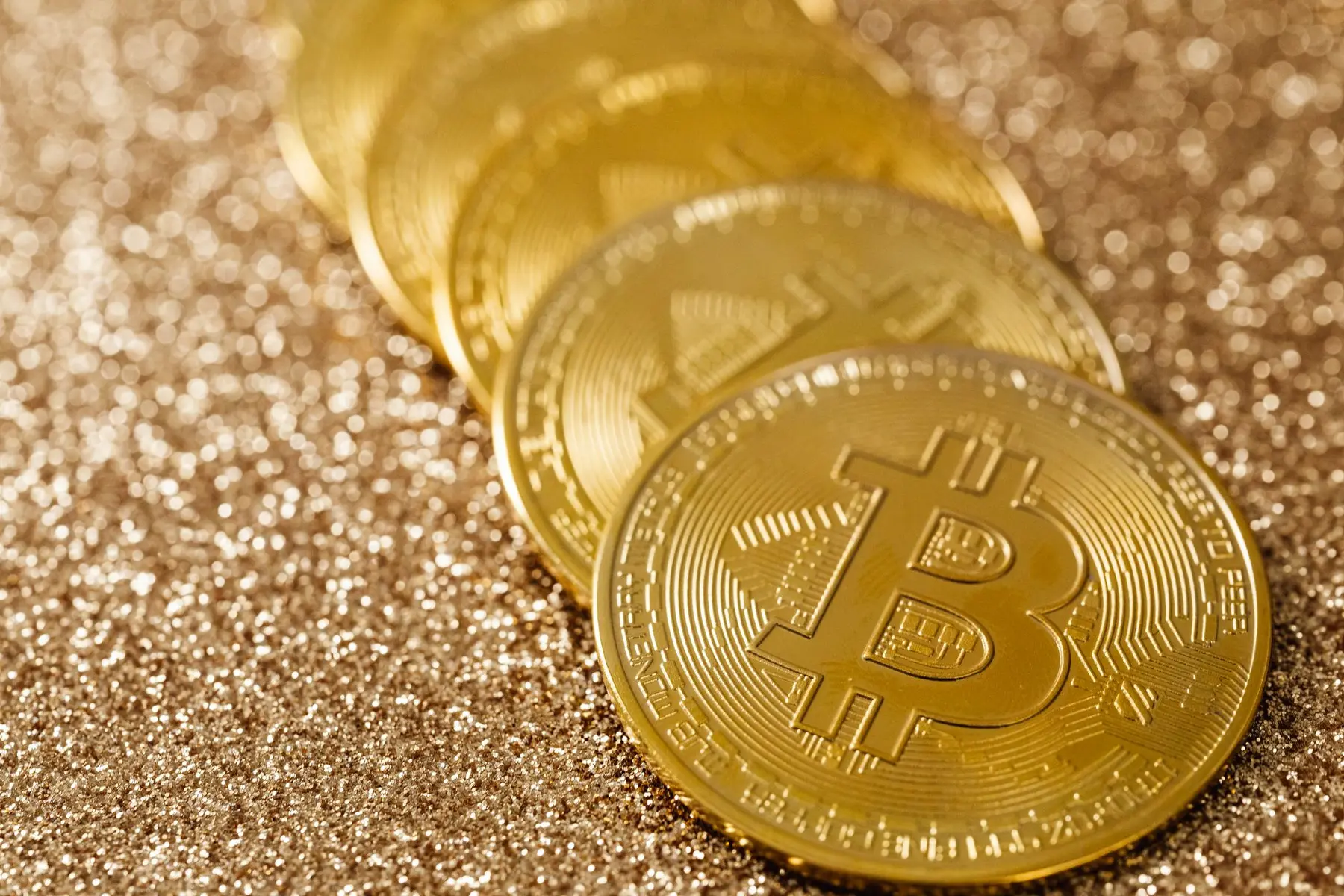 A row of gold-colored Bitcoin physical tokens resting on a shimmering gold glitter surface