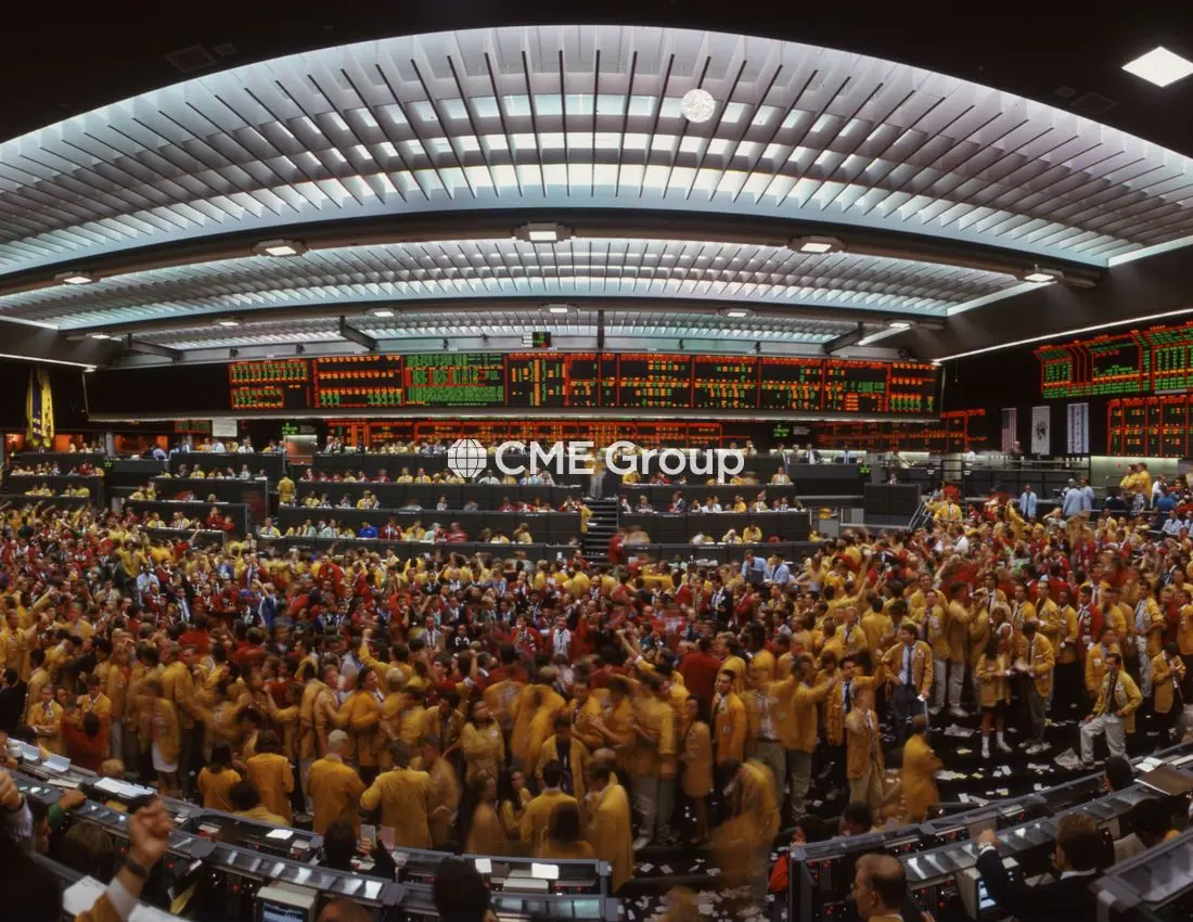 Wide shot of traders in colorful jackets on the crowded CME Group trading floor with digital price boards.