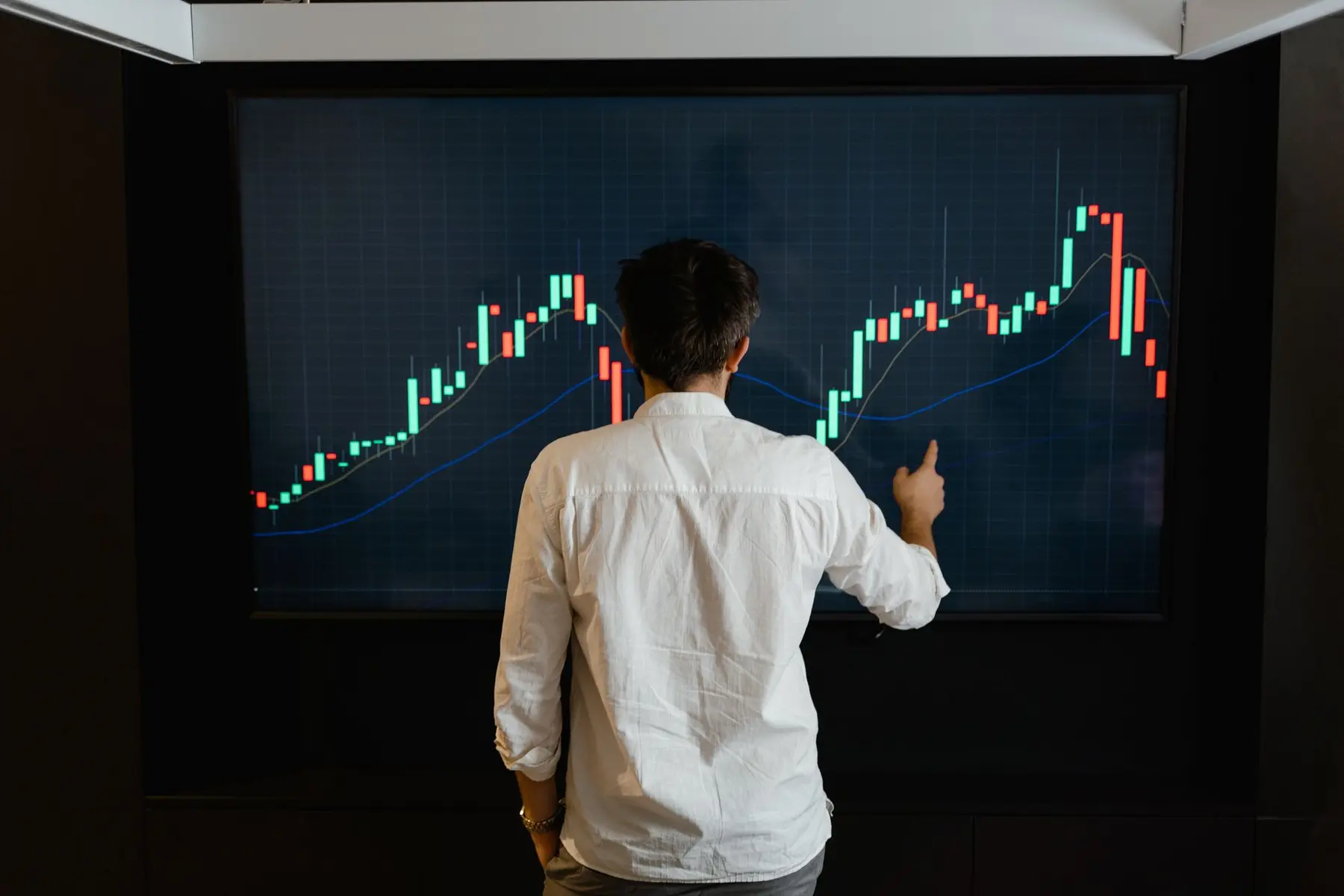 Back view of a man in a white shirt pointing at a large screen displaying financial candlestick charts