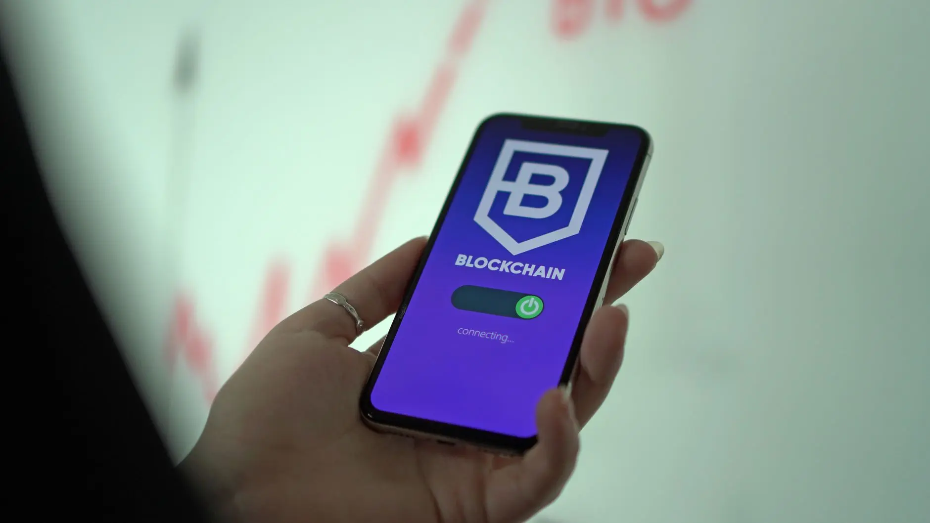 A hand holding a smartphone displaying a Blockchain app interface with a purple background and a power button.