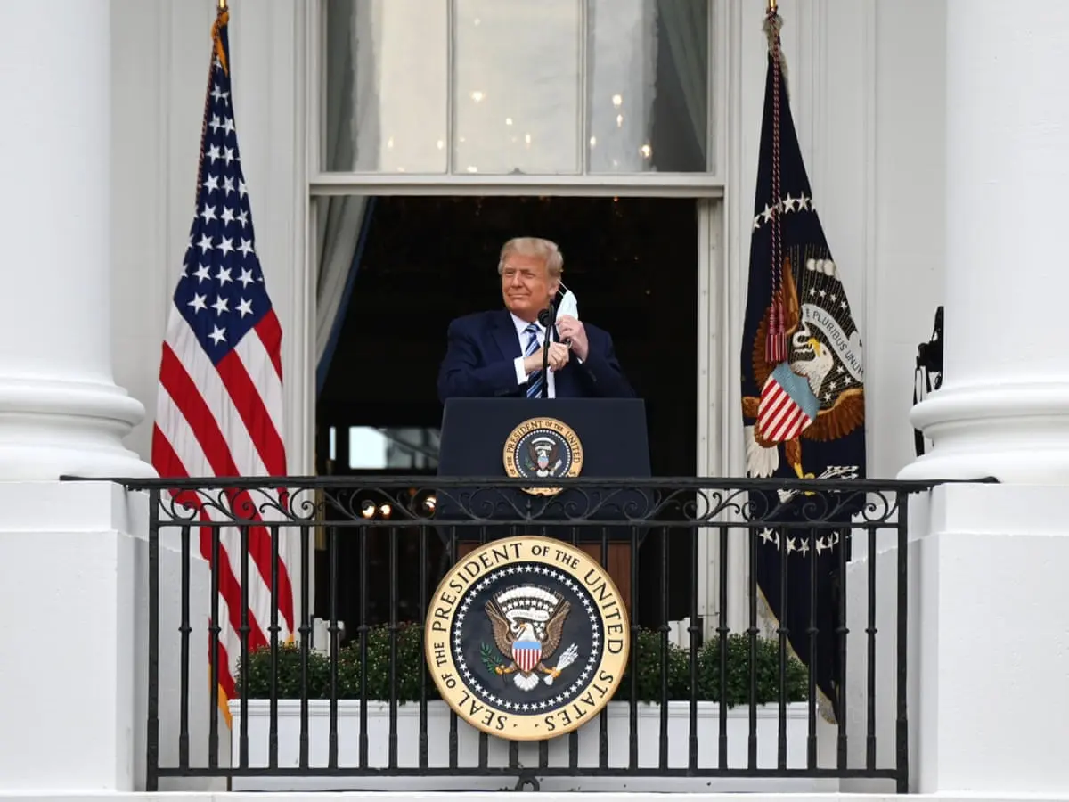 Donald Trump speaking at a podium on a White House balcony, flanked by American and Presidential flags.