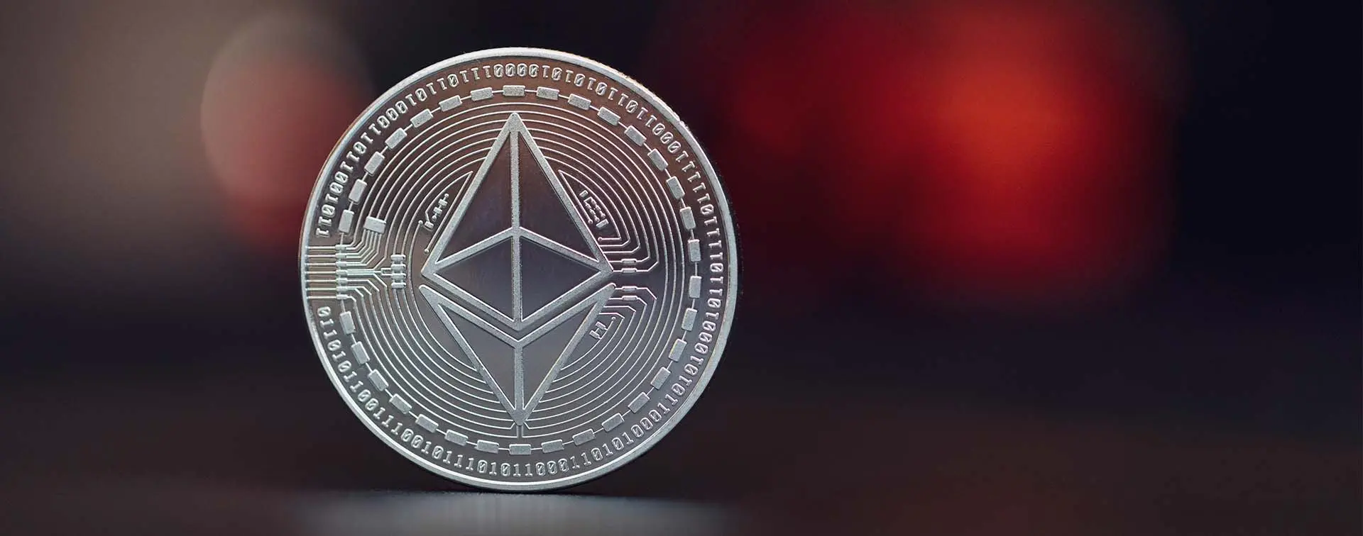 Close-up of a silver physical Ethereum coin with the diamond-shaped logo against a blurred dark red background