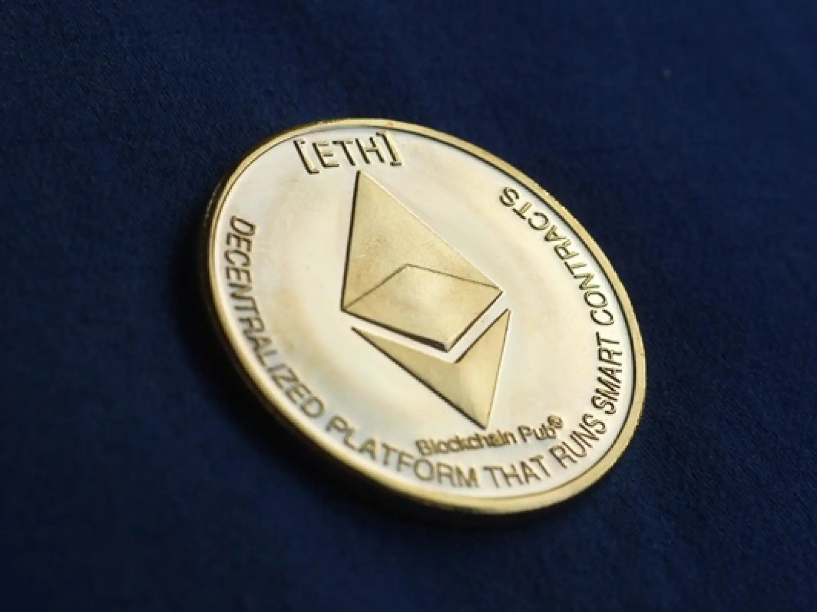 Gold-colored physical Ethereum coin with ETH symbol and text on a dark blue background