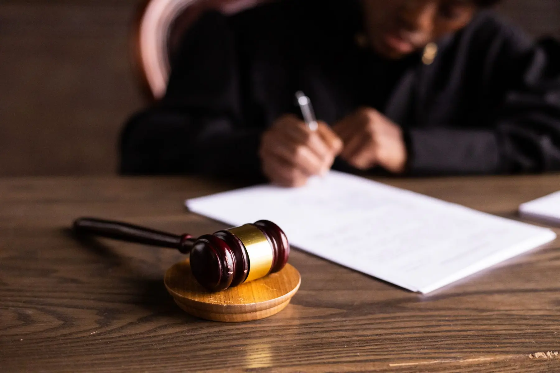 A wooden judge's gavel on a sound block next to official documents and a person writing with a pen.