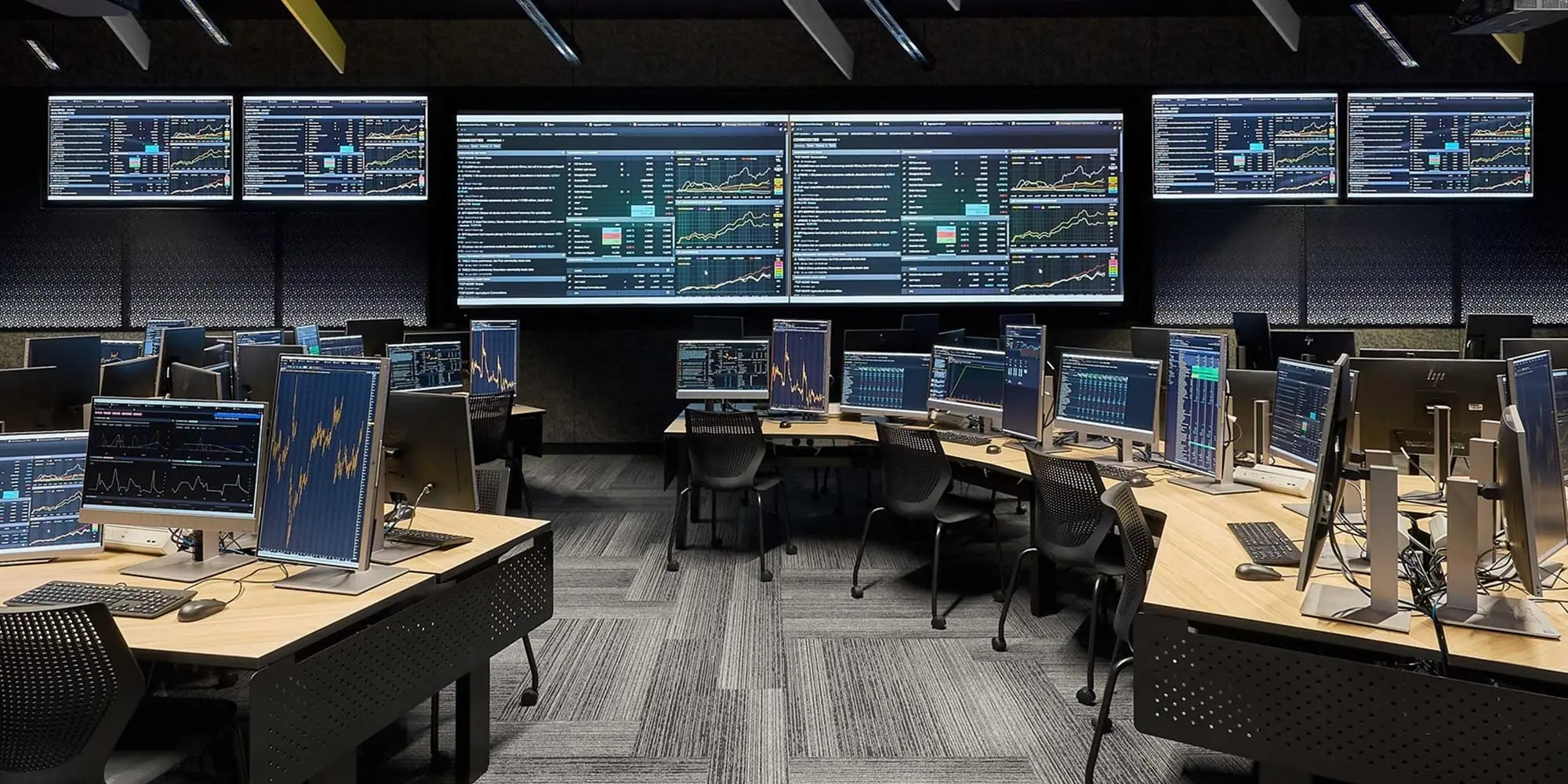 A professional institutional trading room with multiple workstations and large wall-mounted data monitors.