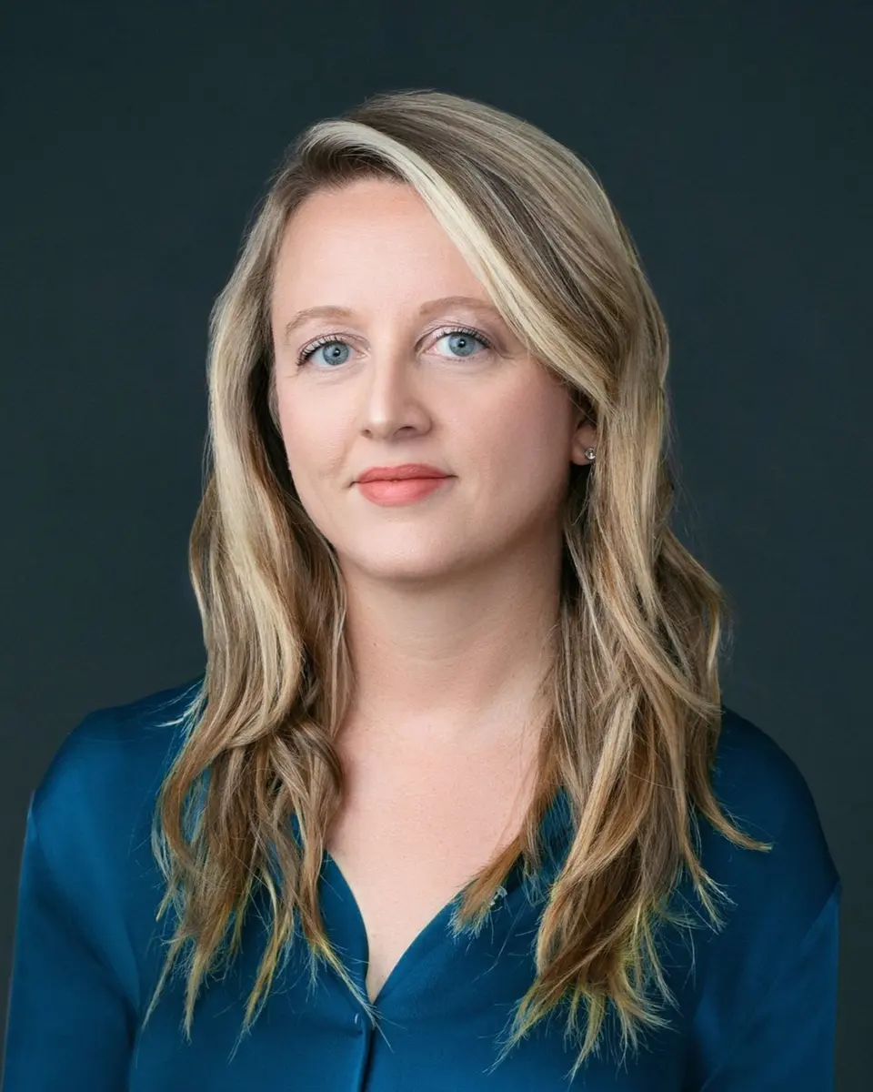 Headshot of Janine Yorio with blonde hair wearing a blue satin blouse against a dark grey background