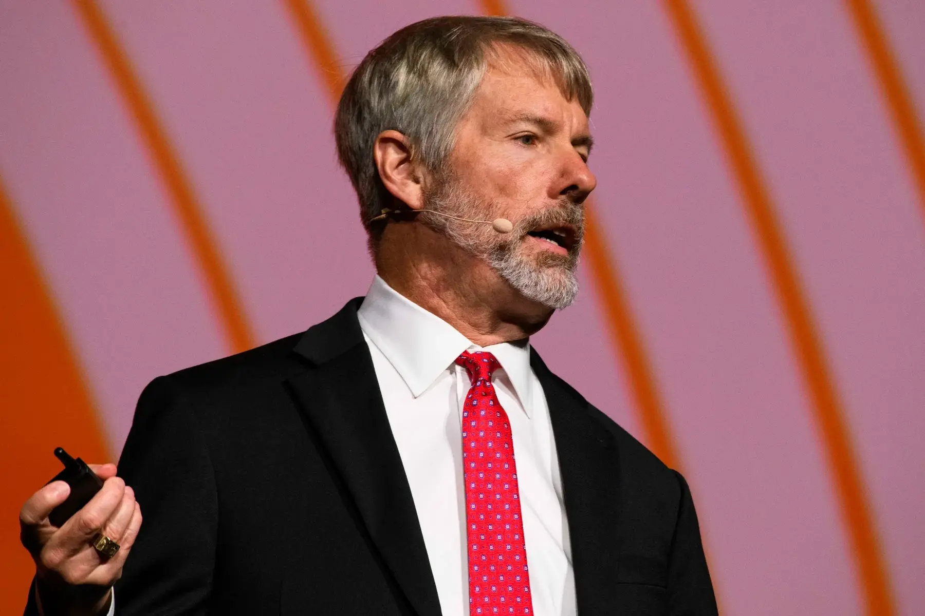 Michael Saylor speaking at a conference, wearing a black suit and red tie with a headset microphone.