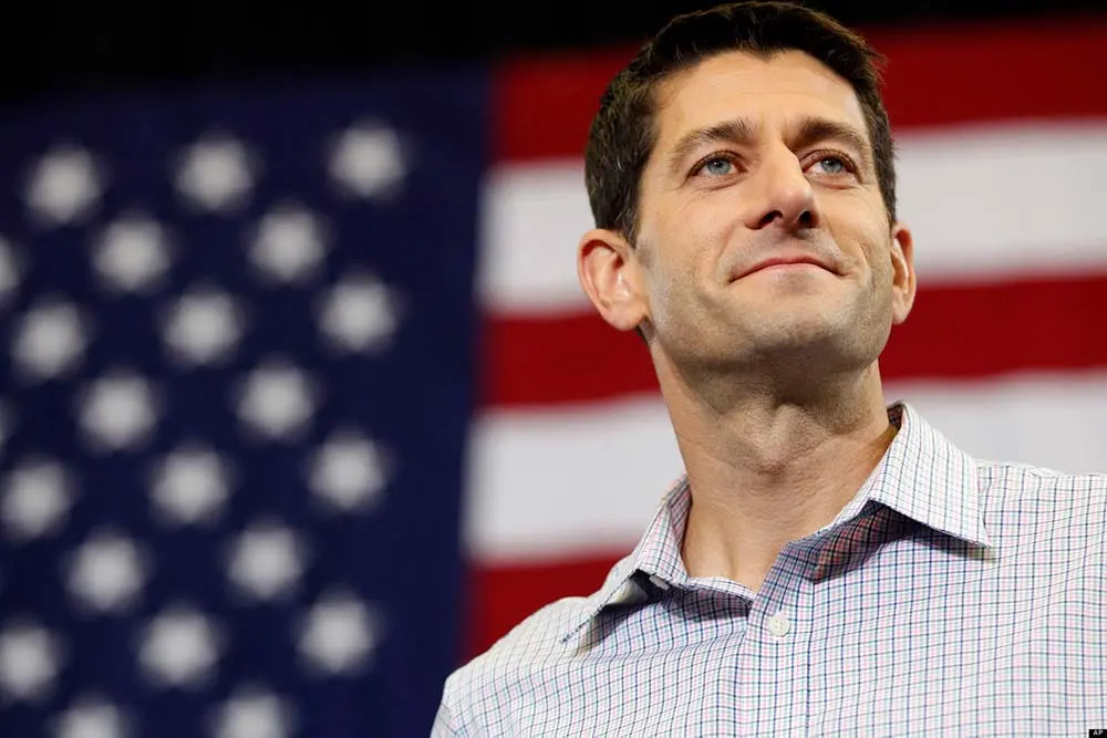 Paul Ryan wearing a checkered shirt and smiling in front of a blurred American flag.