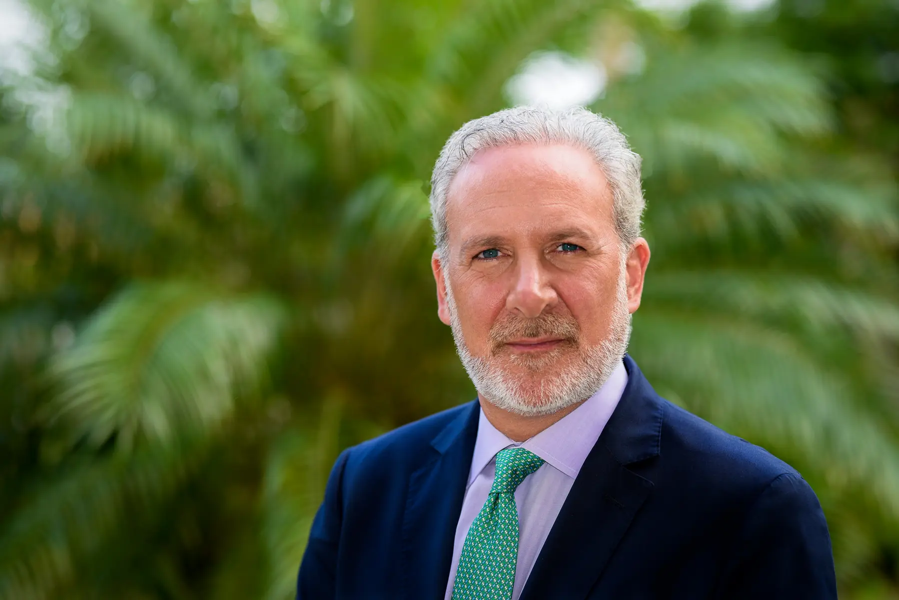 Portrait of Peter Schiff wearing a navy blue suit, purple shirt, and green patterned tie against a lush green foliage background.