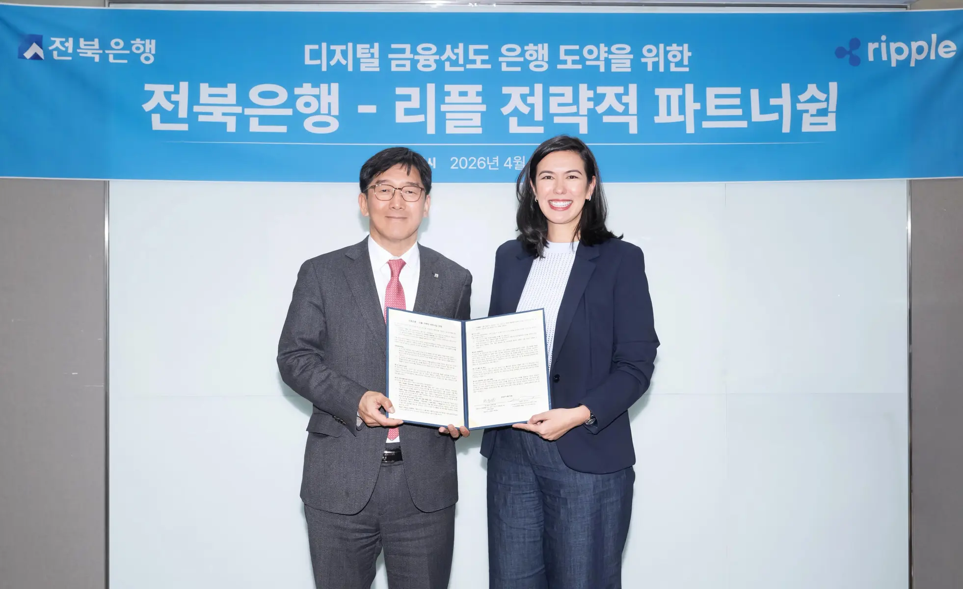 Two professionals holding a signed partnership agreement in front of a blue banner with Ripple and Jeonbuk Bank logos.