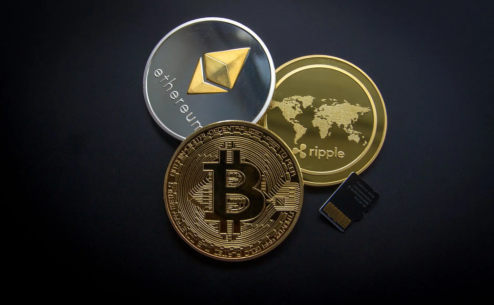 Physical coins representing Bitcoin, Ethereum, and Ripple on a black background with a small microchip.