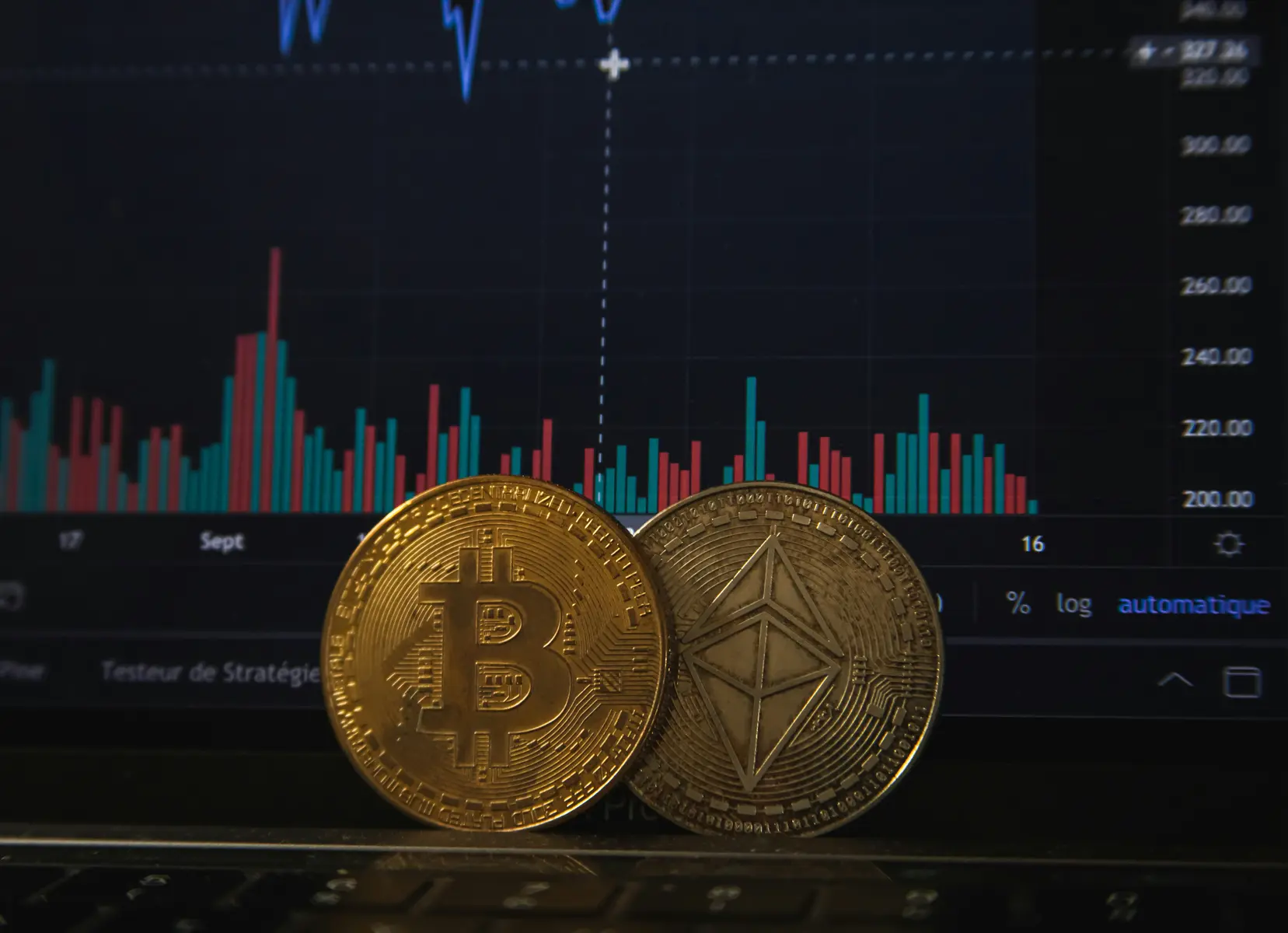 Physical Bitcoin and Ethereum coins in front of a laptop screen displaying a cryptocurrency trading chart.