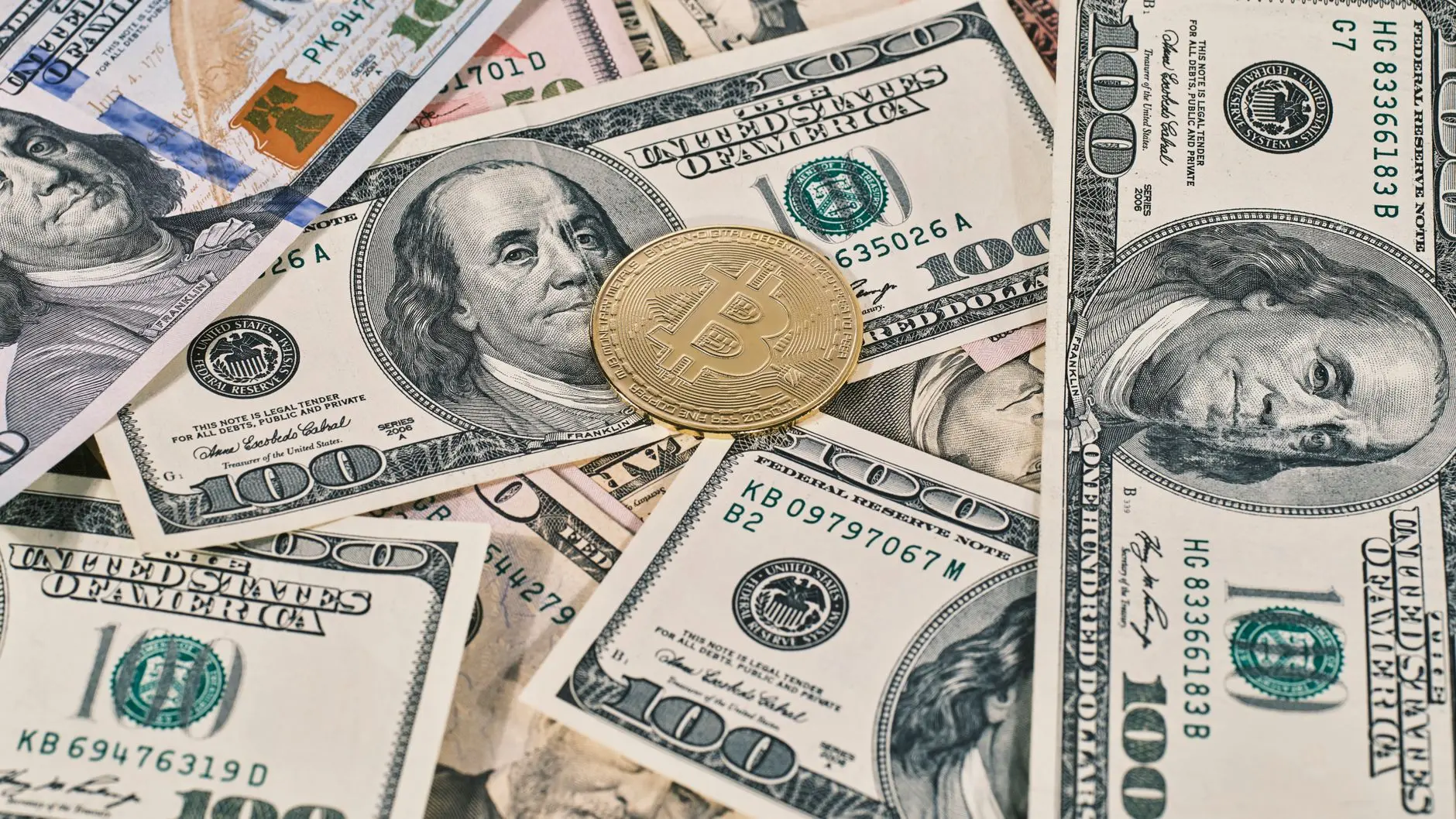 A gold Bitcoin physical coin resting on a scattered pile of US one hundred dollar bills