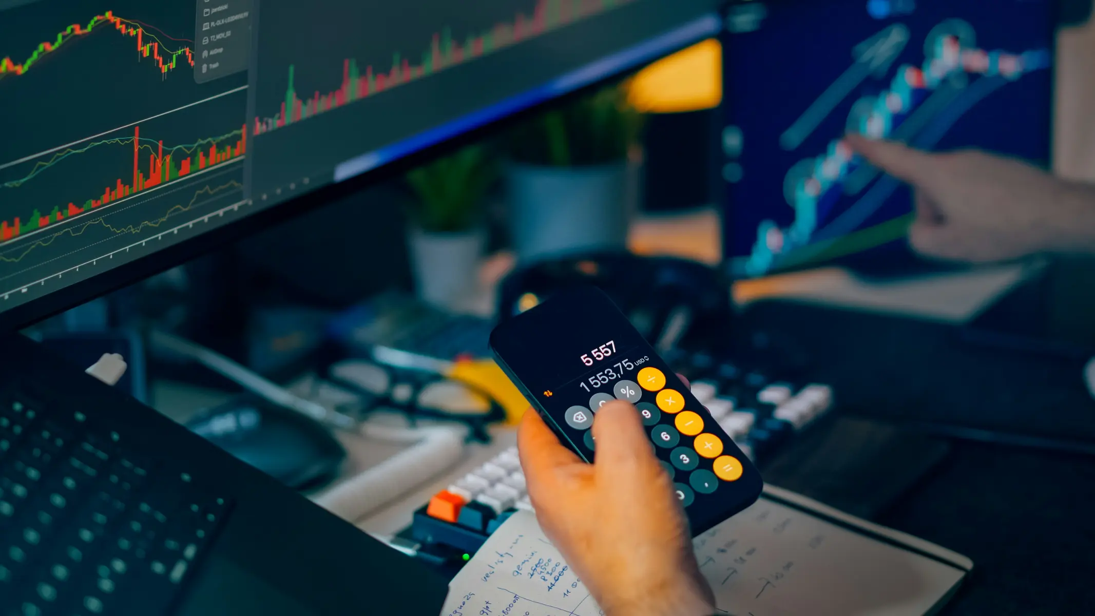 A person using a smartphone calculator in front of computer monitors displaying stock market candlestick charts.