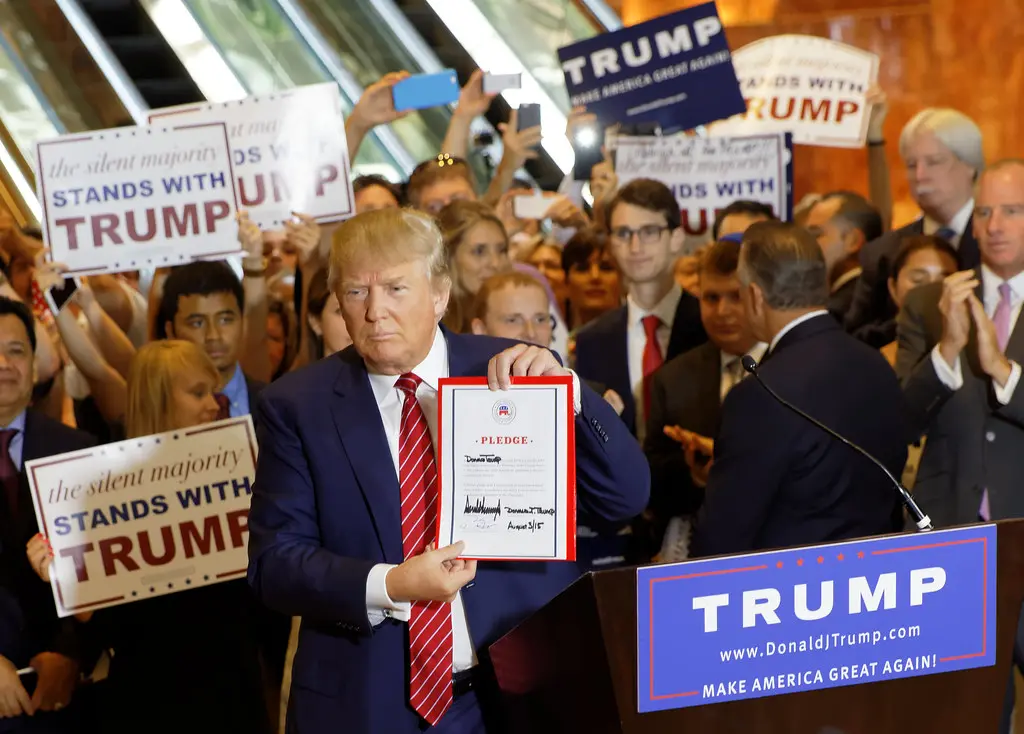 Donald Trump holds up a signed pledge document while standing behind a campaign podium.