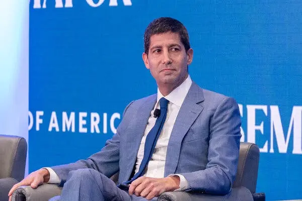 Kevin Warsh wearing a blue suit and tie, seated in front of a blue backdrop with the words 'OF AMERICA'.