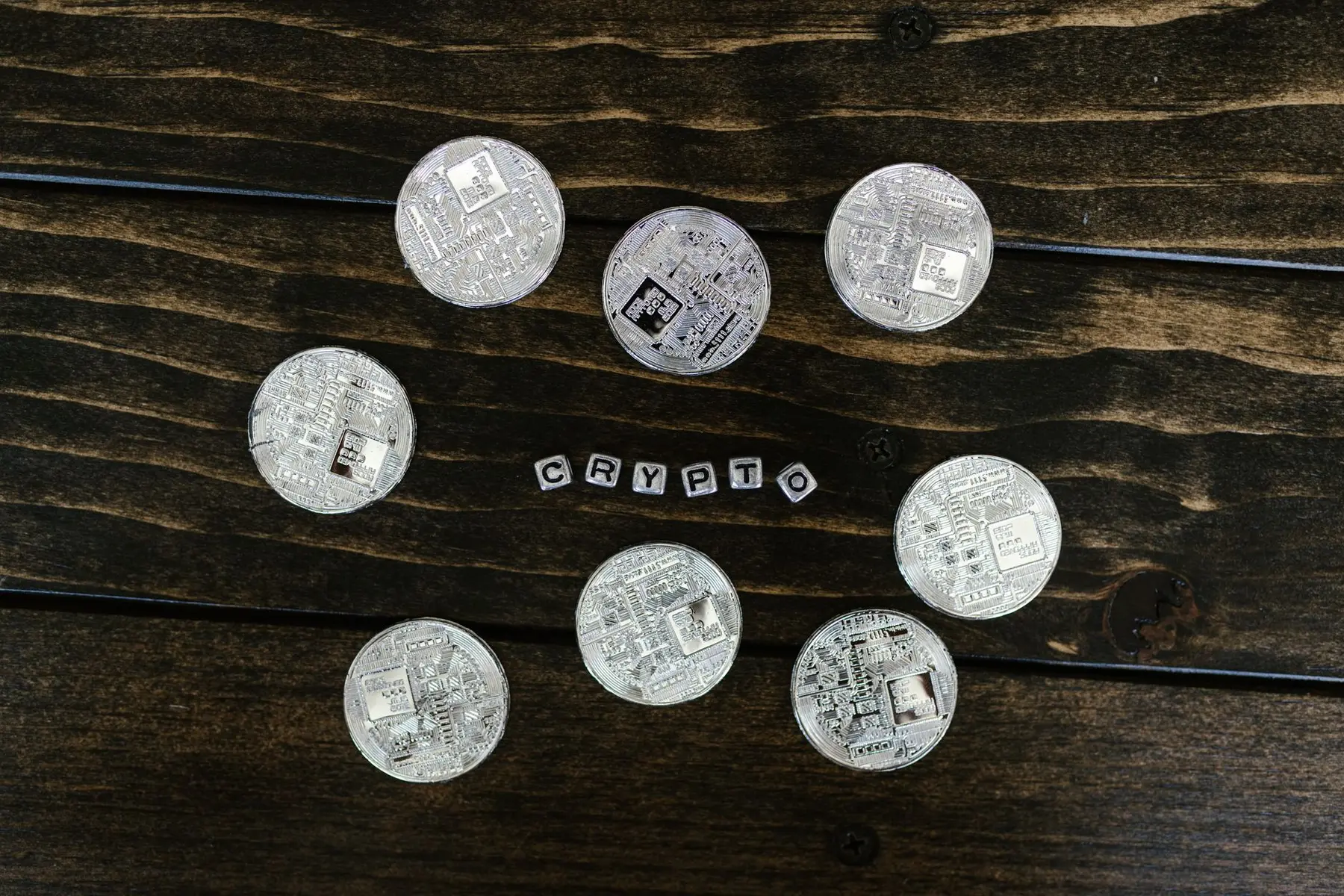 Physical silver-colored cryptocurrency coins surrounding the word CRYPTO in small block letters on a dark wood surface