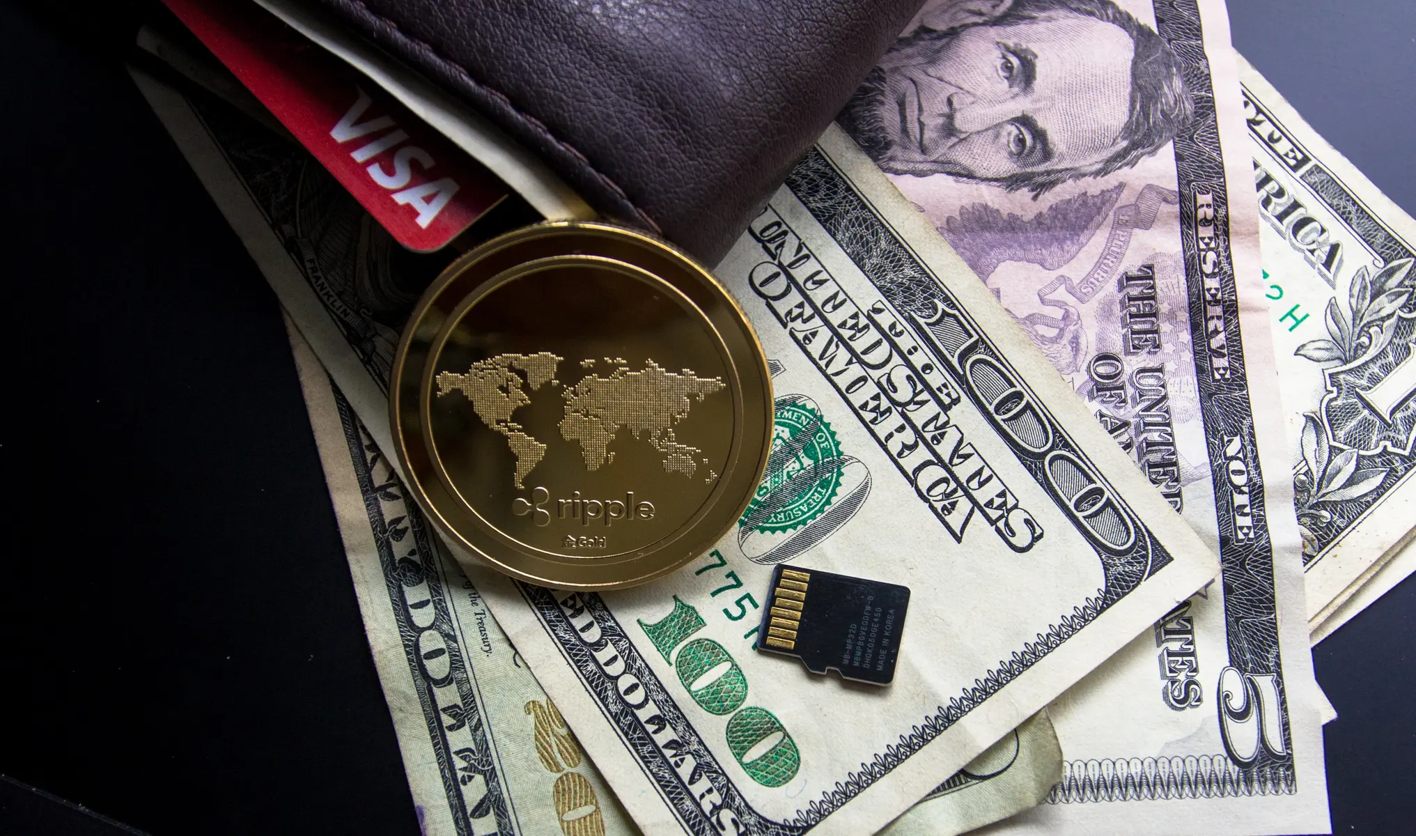 A gold Ripple XRP coin resting on US dollar bills next to a leather wallet and a memory card.